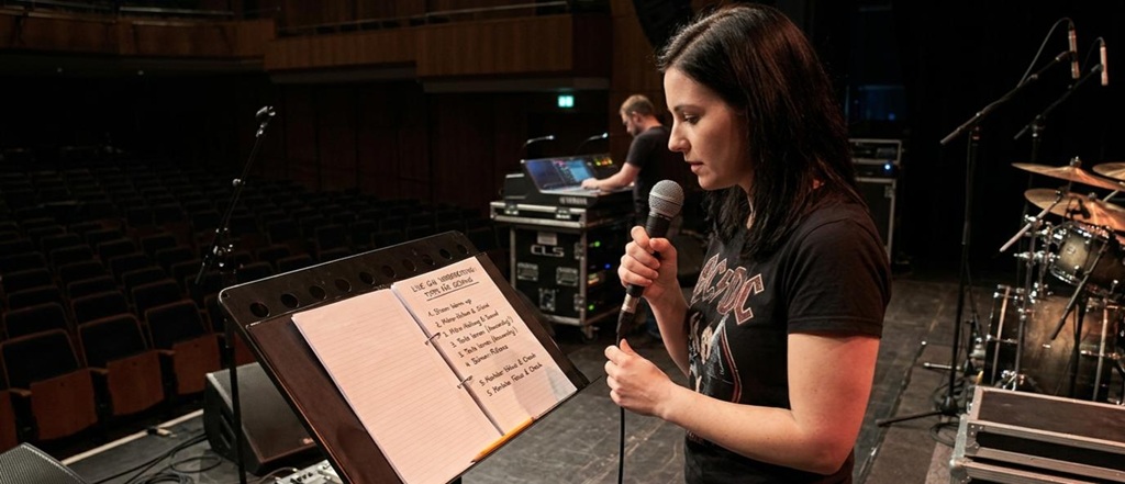 Eine Sängerin steht auf einer Bühne während eines Soundchecks, hält ein Mikrofon und liest eine handschriftliche Checkliste in einem Notizbuch. Die Liste im Notizbuch enthält Tipps zur Gesangs-Live-Gig-Vorbereitung, darunter Stimmen-Aufwärmen, Textüben und Technik-Checks. Ein Tontechniker ist im Hintergrund an einem Mischpult zu sehen.