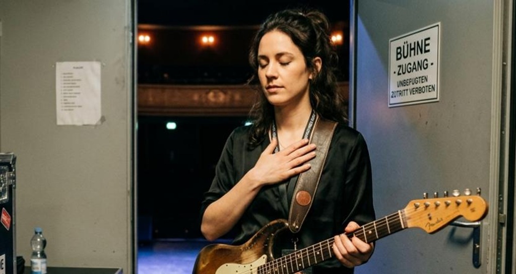 Eine Musikerin steht mit geschlossenen Augen und der Hand auf der Brust im Backstage-Bereich, um vor ihrem Auftritt tief durchzuatmen. Sie hält eine E-Gitarre. Im Hintergrund öffnet sich die Tür zur abgedunkelten Konzertbühne und dem Zuschauersaal. Links steht ein Flightcase mit einem „sound-check.de“-Aufkleber, darüber hängt ein Zettel mit der Aufschrift „Routine: Tief atmen, Fokus, Lächeln“.