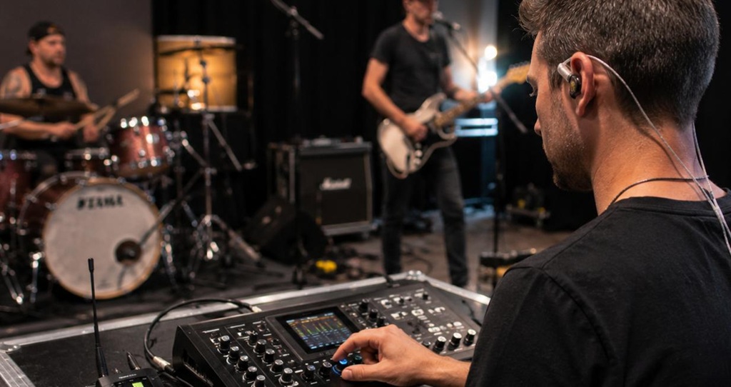 Musiker mit In-Ear-Kopfhörern stellt im Proberaum an einem Digitalmixer seinen Monitor-Mix ein, während Schlagzeuger und Gitarrist im Hintergrund proben.