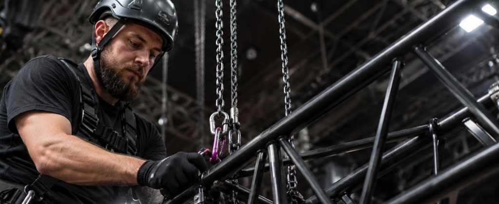 Rigging-Techniker mit Helm und Sicherheitsausrüstung sichert eine geflogene Traverse mit Ketten und Karabinern in einer Veranstaltungshalle.
