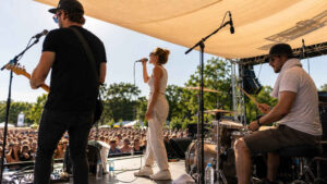 Band spielt bei einem sonnigen Open-Air-Gig auf einer Bühne mit Sonnensegel, Wasserflaschen und Publikum im Hintergrund.