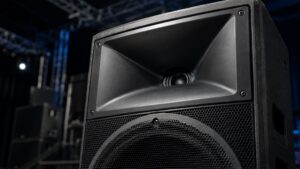 Nahaufnahme eines PA-Lautsprechers mit deutlich sichtbarem Waveguide im Hochtonbereich vor unscharfer Bühnenumgebung.